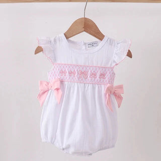 A white baby romper with pink embroidered trim, pink bows, and ruffled sleeves, hanging on a wooden hanger against a plain light background.