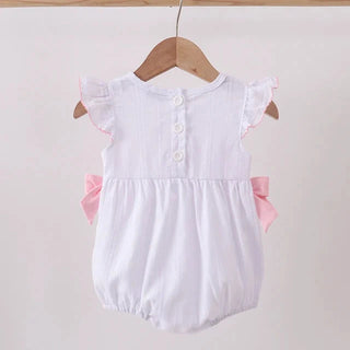 A white baby romper with pink ruffle accents and side bows, hanging on a wooden hanger from a light wood clothing rack against a plain off-white wall.