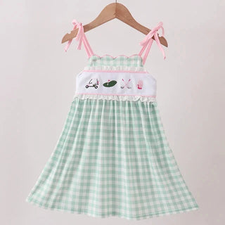 A light green and white gingham dress with pink shoulder straps and a white chest panel featuring golf-themed illustrations, hanging on a wooden hanger against a light pink background.
