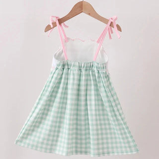 A light green and white gingham pinafore dress with pink shoulder ties, hanging on a wooden hanger against a plain light background.