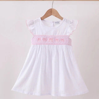 A white girl's dress with a pink smocked chest band and ruffle sleeves, hanging on a wooden hanger against a plain light background.