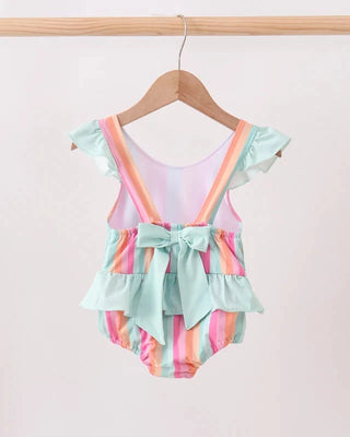 A toddler girl's ruffled swimsuit hanging on a wooden hanger against a light pink background. The swimsuit features pastel mint green ruffles, pastel rainbow stripes (pink, orange, yellow, green, blue), and a mint green bow at the center of the waist.