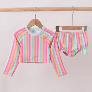 A two-piece children's swim set hanging on wooden hangers from a light wood rack. The set includes a long-sleeve cropped top and matching bloomers, both featuring pastel vertical stripes in pink, orange, yellow, green, and blue on a white base.
