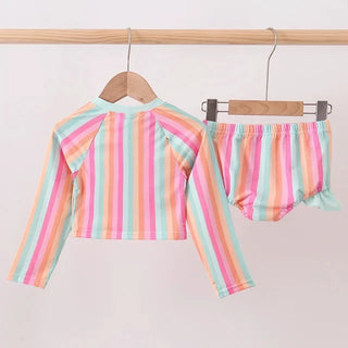 A two-piece baby swim  set hanging on wooden hangers from a light wood rail. The set includes a long-sleeve cropped top and matching bloomers, both featuring pastel vertical stripes in pink, orange, yellow, green, and blue on a white base.