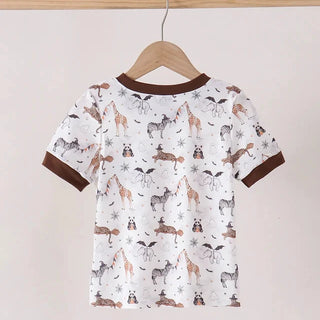 A children's short-sleeve t-shirt hanging on a wooden hanger against a plain light beige wall. The shirt has a white base with a woodland animal print featuring brown, black, and gray foxes, deer, and birds, with contrasting dark brown ribbed collar and sleeve cuffs.