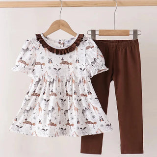 A toddler girl's two-piece clothing set hanging on wooden hangers against a plain beige wall. The set includes a white short-sleeve ruffled blouse with a brown collar and an all-over woodland animal print, paired with solid brown leggings.