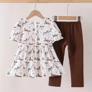 A toddler girl's two-piece outfit hanging on wooden hangers against a plain beige wall. The set includes a white short-sleeve ruffled blouse with an all-over animal print in brown, black, and gray, paired with solid dark brown leggings.