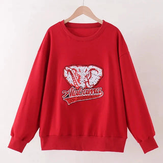 A red long-sleeve crewneck sweatshirt hanging on a wooden hanger against a plain off-white wall. The sweatshirt features a front graphic of the Alabama Crimson Tide ram logo in red and white, with 'Alabama Crimson Tide' written in script below.