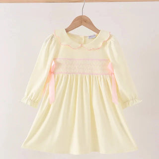 A pale yellow long-sleeve dress with a pink smocked chest panel and matching pink ribbon bows, hanging on a wooden hanger against a plain light background.