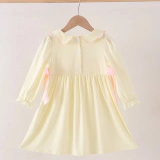 A pale yellow long-sleeve dress with a Peter Pan collar, buttoned neckline, and pink ribbon accents on the sides, hanging on a wooden hanger from a wooden rod against a plain light background.