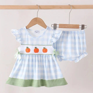 A baby girl's two-piece outfit set consisting of a light blue and white gingham dress with a green hem, three orange embroidered pumpkins on the chest, and matching ruffled shorts, hanging on wooden hangers against a light beige background.