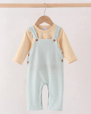 Baby romper set hanging on a wooden hanger, featuring a light sage green and white diamond patterned overall jumpsuit over a short-sleeve cream shirt.
