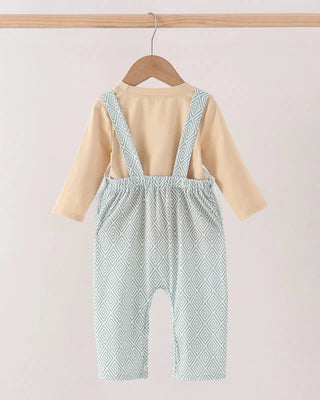 A baby romper set hanging on a wooden hanger from a wooden rod. The outfit features a light beige long-sleeve top layered under sage green patterned overall-style pants with shoulder straps.
