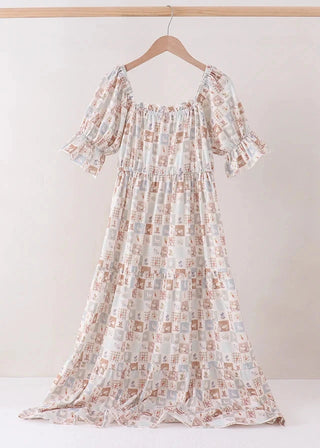 A long white maxi dress with a brown and beige block-print pattern, featuring puff sleeves and a smocked bodice, hanging on a wooden hanger against a neutral beige background.