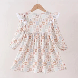 A long-sleeve dress with a patchwork print in muted brown, beige, and light blue, hanging on a wooden hanger against a plain off-white wall.