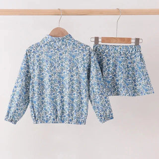 A matching two-piece clothing set featuring a long-sleeve cropped jacket and a high-waisted mini skirt, both in a light blue and white floral print, hanging on wooden hangers against a plain off-white wall.