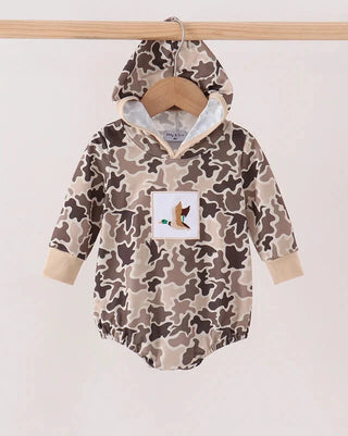 A baby hooded romper in a beige, brown, and gray camouflage pattern, hanging on a wooden hanger from a light wood rack against a plain off-white background. The romper features a white square patch on the chest with a cartoon fox illustration.