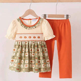 A toddler girl's two-piece outfit hanging on wooden hangers against a plain light background. The set includes a short-sleeve dress with a cream gingham upper bodice, a smocked orange-trimmed chest band with pumpkin motifs, and a green skirt printed with pumpkins and acorns, paired with solid bright orange leggings.