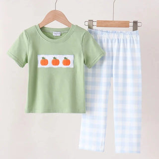 A children's two-piece outfit set hanging on wooden hangers against a light beige wall. The set includes a light sage green short-sleeve top with a white rectangular chest patch featuring three orange pumpkins, and light blue and white gingham check pajama pants.