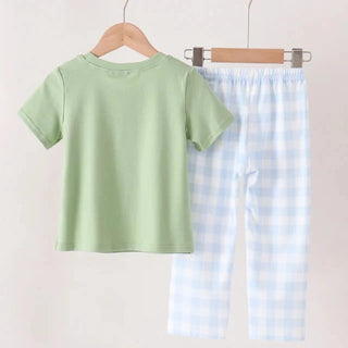 A two-piece children's outfit hanging on wooden hangers against a plain light beige wall. The set includes a light sage green short-sleeve t-shirt and light blue and white gingham checkered pants.