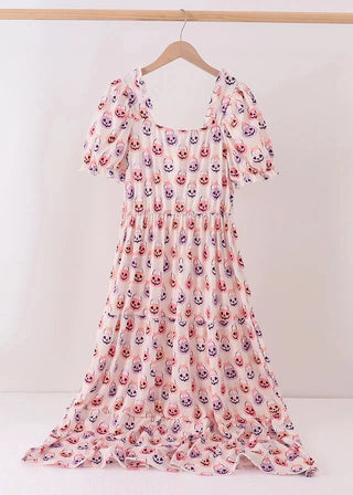 A white midi dress with a repeating pattern of smiling cartoon faces in pink, purple, and orange, featuring puff sleeves and a square neckline, hanging on a wooden hanger against a white wall.