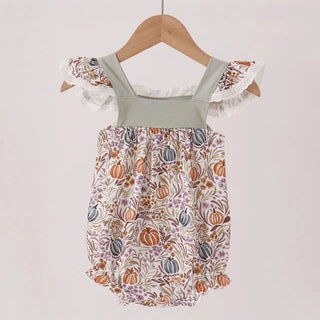 A baby romper with a sage green solid shoulder yoke and ruffled off-the-shoulder sleeves, paired with a cream-colored bodice printed with a fall harvest pattern of pumpkins, gourds, and leaves. The romper is hanging on a wooden hanger from a light wood clothes rack against a plain, light beige wall.
