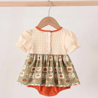 A baby girl's short-sleeve romper dress hanging on a wooden hanger. The top half features a warm cream and orange gingham check pattern, while the bottom half is a sage green skirt with a pumpkin print.
