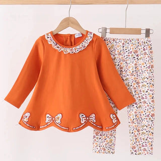 A toddler girl's two-piece clothing set hanging on wooden hangers against a white background. The set includes a bright orange long-sleeve top with a white floral lace collar and embroidered bow details at the hem, paired with matching orange and white floral print pants.