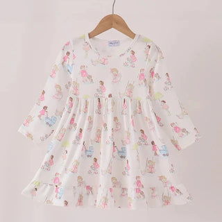 A white long-sleeve baby dress with a colorful cartoon girl print, hanging on a wooden hanger from a light wood rod against a pale pink background.