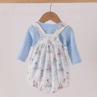 A baby romper set featuring a light blue long-sleeve shirt and white overalls with a colorful bicycle print, hanging on a wooden hanger from a wooden rod against a light beige background.