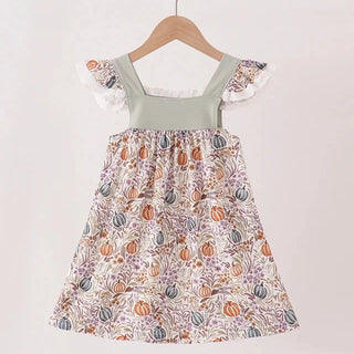 A toddler girl's sleeveless dress with ruffled cap sleeves, featuring a sage green smocked bodice and a white skirt printed with orange, purple, and gray pumpkins and autumn leaves. The dress is hanging on a wooden hanger from a light wood rod against a plain off-white wall.