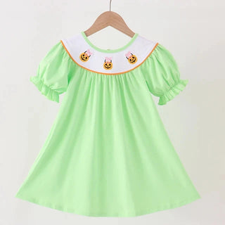 A light green short-sleeve girl dress hanging on a wooden hanger against a white wall. The dress features a white Peter Pan collar decorated with embroidered cartoon faces and a thin orange trim.