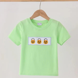 A light green short-sleeve t-shirt hanging on a wooden hanger against a plain off-white wall. The shirt features a rectangular chest graphic containing three embroidered orange jack-o'-lantern faces inside black-outlined lock graphics.