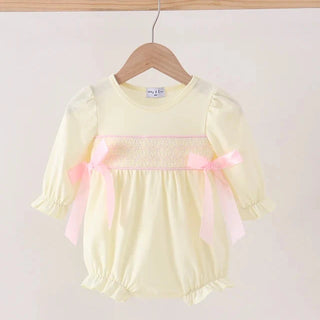 A pale yellow baby romper with pink smocked detailing and pink ribbon bows, hanging on a wooden hanger against a light beige background.