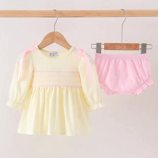 A baby girl's two-piece outfit set consisting of a pale yellow long-sleeve smocked dress with pink shoulder bows and matching light pink ruffled bloomers, displayed on wooden hangers hanging from a wooden rod against a plain light background.