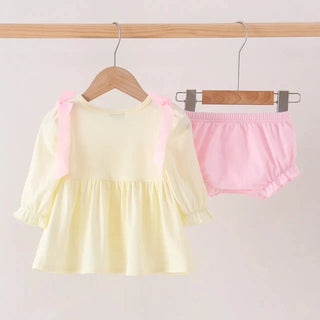 A baby girl's two-piece outfit hanging on wooden hangers against a light beige wall. The set includes a pale yellow long-sleeve blouse with pink shoulder ties and matching light pink ruffled bloomers.