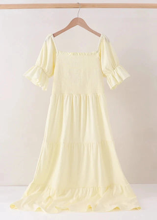 A pale yellow tiered dress with ruffled short sleeves and a smocked bodice, hanging on a wooden hanger against a plain light beige background.