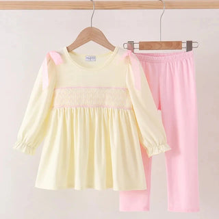 A two-piece clothing set hanging on wooden hangers against a light beige wall. The set includes a pale yellow long-sleeve smocked blouse with pink shoulder straps and a pink smocked chest band, paired with pink pants.