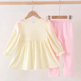 A girl's two-piece clothing set hanging on wooden hangers against a light pink background. The set includes a pale yellow long-sleeve tunic dress with ruffled sleeves and a matching pair of light pink pants.