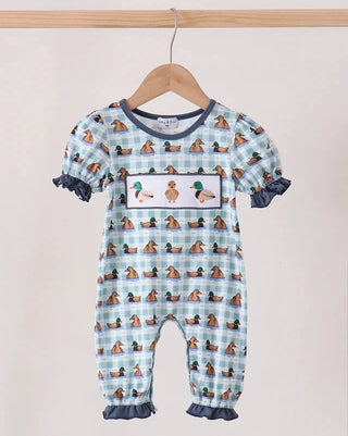 A baby short-sleeve romper hanging on a wooden hanger against a light beige wall. The romper features a light blue and white gingham pattern with a repeating print of ducks, a central chest patch showing a duck and a green hat, and dark blue ruffled trim on the sleeves, cuffs, and leg hems.