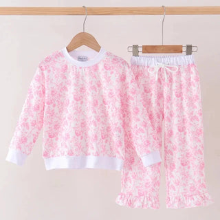 A two-piece children's pants set hanging on wooden hangers against a plain light beige wall. The set includes a long-sleeve top and matching pants in a light pink and white floral print, with solid white ribbed trim at the collar, cuffs, and hem.