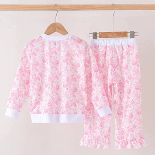 A two-piece children's pants set hanging on wooden hangers against a plain light beige wall. The set includes a long-sleeve top and matching pants in a light pink and white floral print, with solid white ribbed cuffs, hem, and waistband.