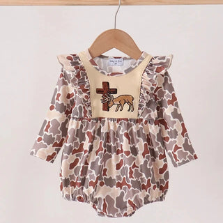 A baby romper with a brown and beige cow print pattern, featuring a solid beige bib-style chest panel with a cross and deer applique, hanging on a wooden hanger.