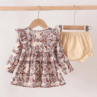A toddler girl's two-piece outfit hanging on wooden hangers from a light wood rack. The set includes a long-sleeve ruffle-shoulder top with a brown, gray, and beige cow print, and matching beige ruffled bloomers.