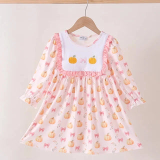 A toddler girl's long-sleeve dress hanging on a wooden hanger against a plain beige wall. The dress is light pink with a white bib-style chest panel featuring two orange pumpkins and pink bows, and is decorated with a scattered pattern of orange pumpkins and pink bows.