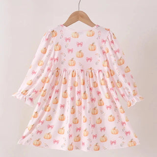A light pink long-sleeve baby dress with an all-over pumpkin and bow pattern, hanging on a wooden hanger against a plain off-white wall.