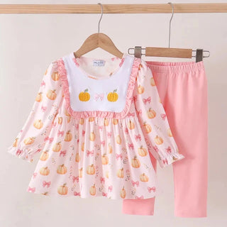 A children's two-piece outfit set hanging on wooden hangers against a plain light background. The set includes a long-sleeve top with a white bib-style chest panel, pink ruffle trim, and an allover pumpkin print, paired with solid pink pants.