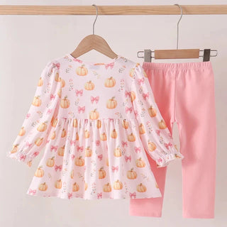 A toddler girl's two-piece clothing set hanging on wooden hangers against a light wall. The set includes a long-sleeve top in white with a pink bow and orange pumpkin print, paired with solid light pink leggings.