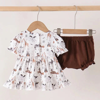 A toddler girl's two-piece outfit hanging on wooden hangers from a light wood rod. The set includes a white short-sleeve dress with a brown, black, and beige woodland animal print, and a pair of solid dark brown bloomers.