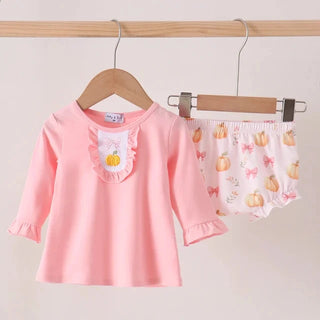 A baby girl's two-piece outfit hanging on wooden hangers against a light beige wall. The set includes a long-sleeve pink top with a ruffled chest panel featuring a small yellow pumpkin applique, and matching pink floral bloomers with a pumpkin and bow pattern.